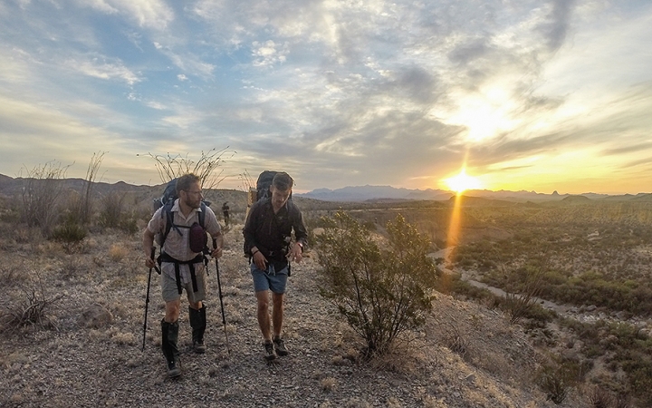 two people hike in a dry landscape while the sun sets behind mountains in the distance